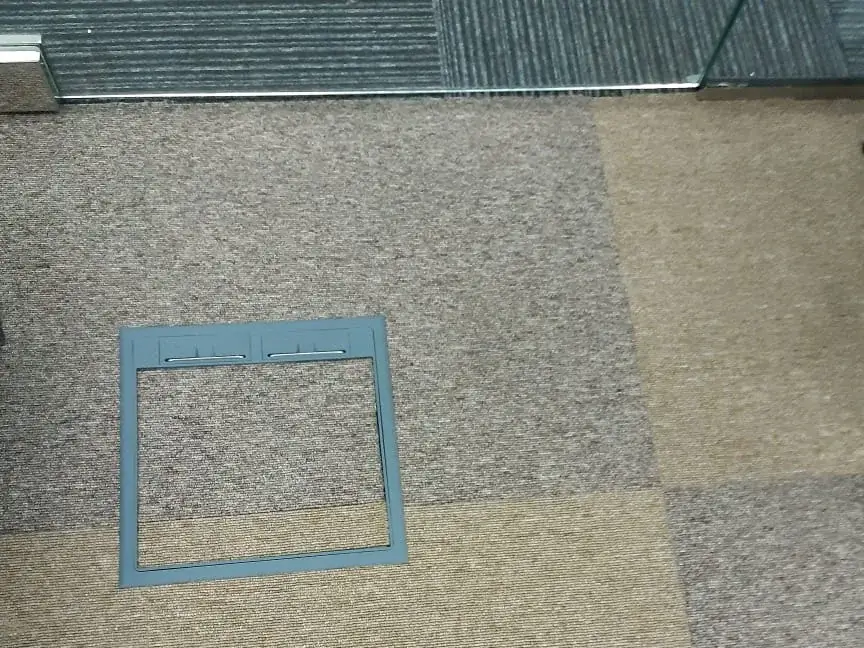 Carpet tile cut around a floor service box, showing the quarter-turn laying and the transition to dark tiles at a glass threshold